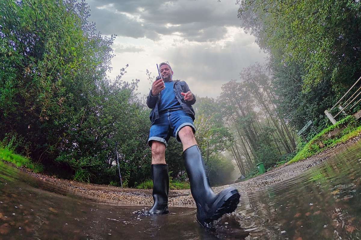 Tom in wellies, holding a radio while filming on a flooded country lane
