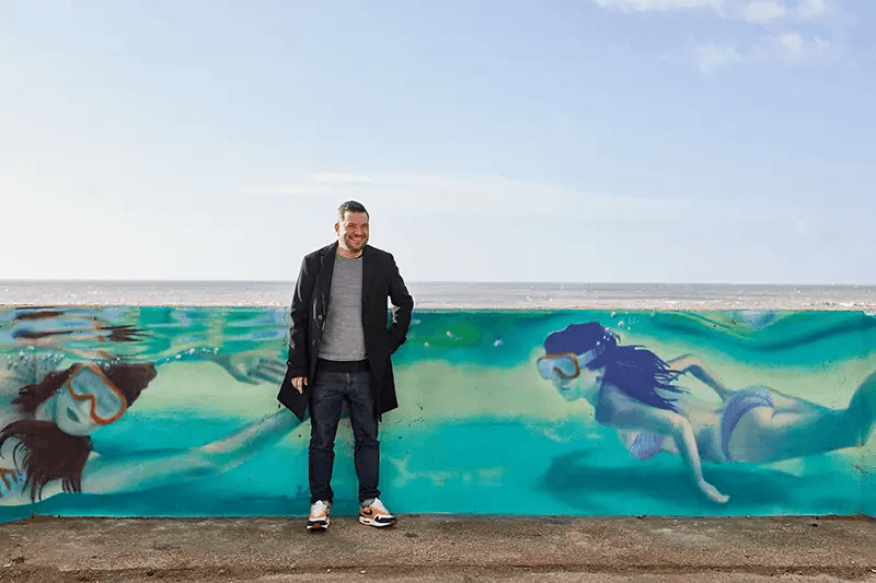 Tom standing by the painted swimmers mural on the seafront sea wall, with the sea behind